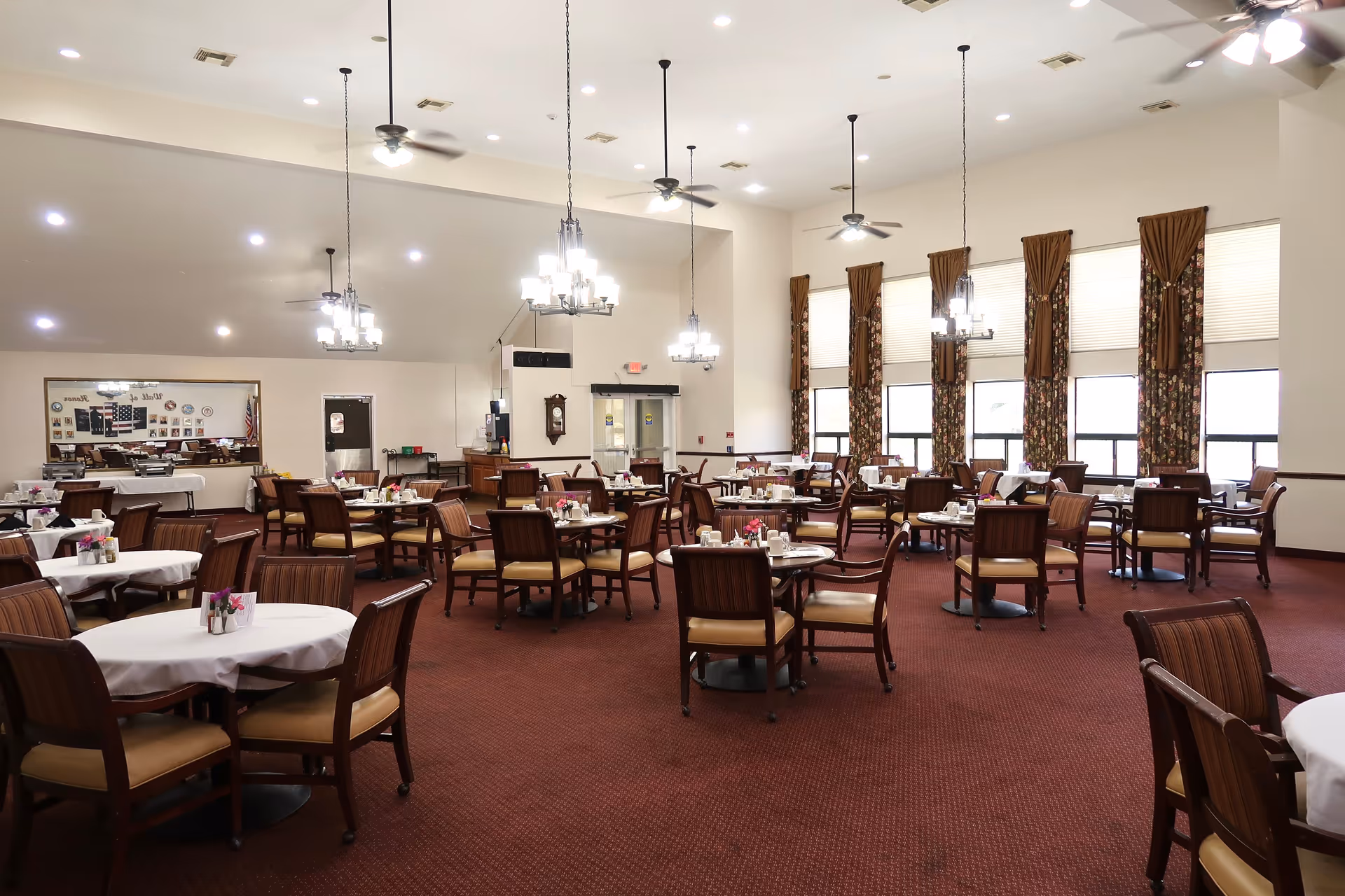 A spacious dining room with multiple round tables covered with white tablecloths, each surrounded by wooden chairs with cushioned seats. The room has high ceilings with ceiling fans and hanging chandeliers. Large windows with floral curtains allow natural light to fill the space. The walls are light-colored, and the carpet is a deep red. There is a large mirror on one wall reflecting part of the room.