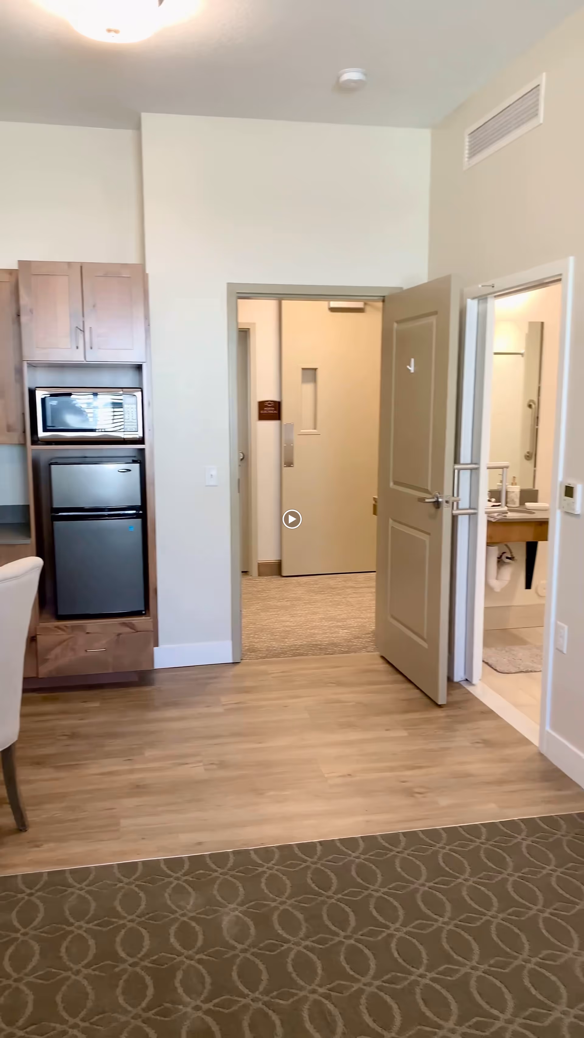 Interior view of a room in Whisper Cove Assisted Living and Memory Care showing a small kitchenette area with a microwave and mini refrigerator built into wooden cabinets on the left. There is a doorway straight ahead leading to a hallway and another open door on the right revealing a bathroom with a sink and mirror. The floor transitions from carpet with a circular pattern to wood laminate.