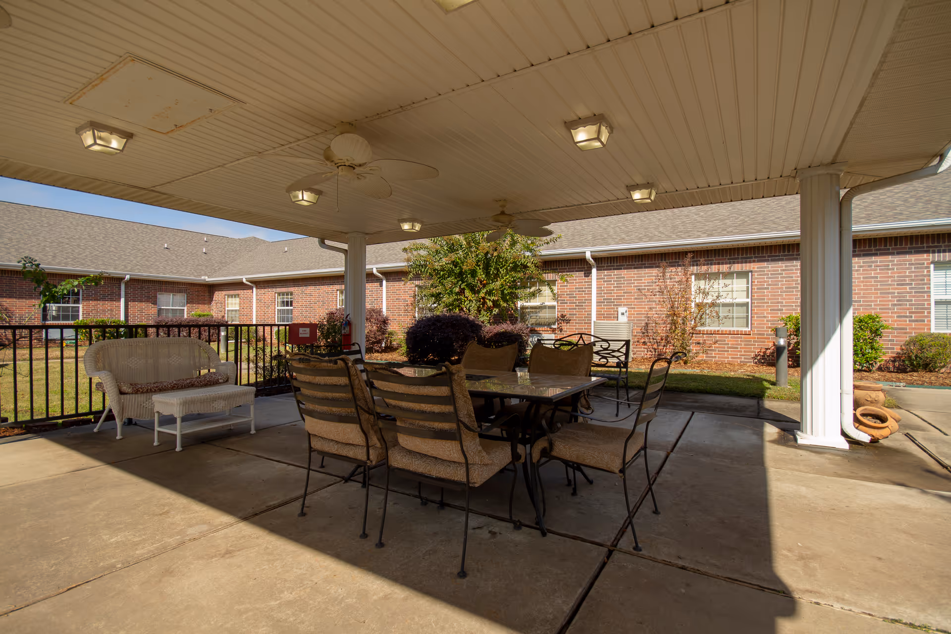 Covered outdoor patio with a dining table and chairs under a roofed pavilion in a courtyard beside a brick building.