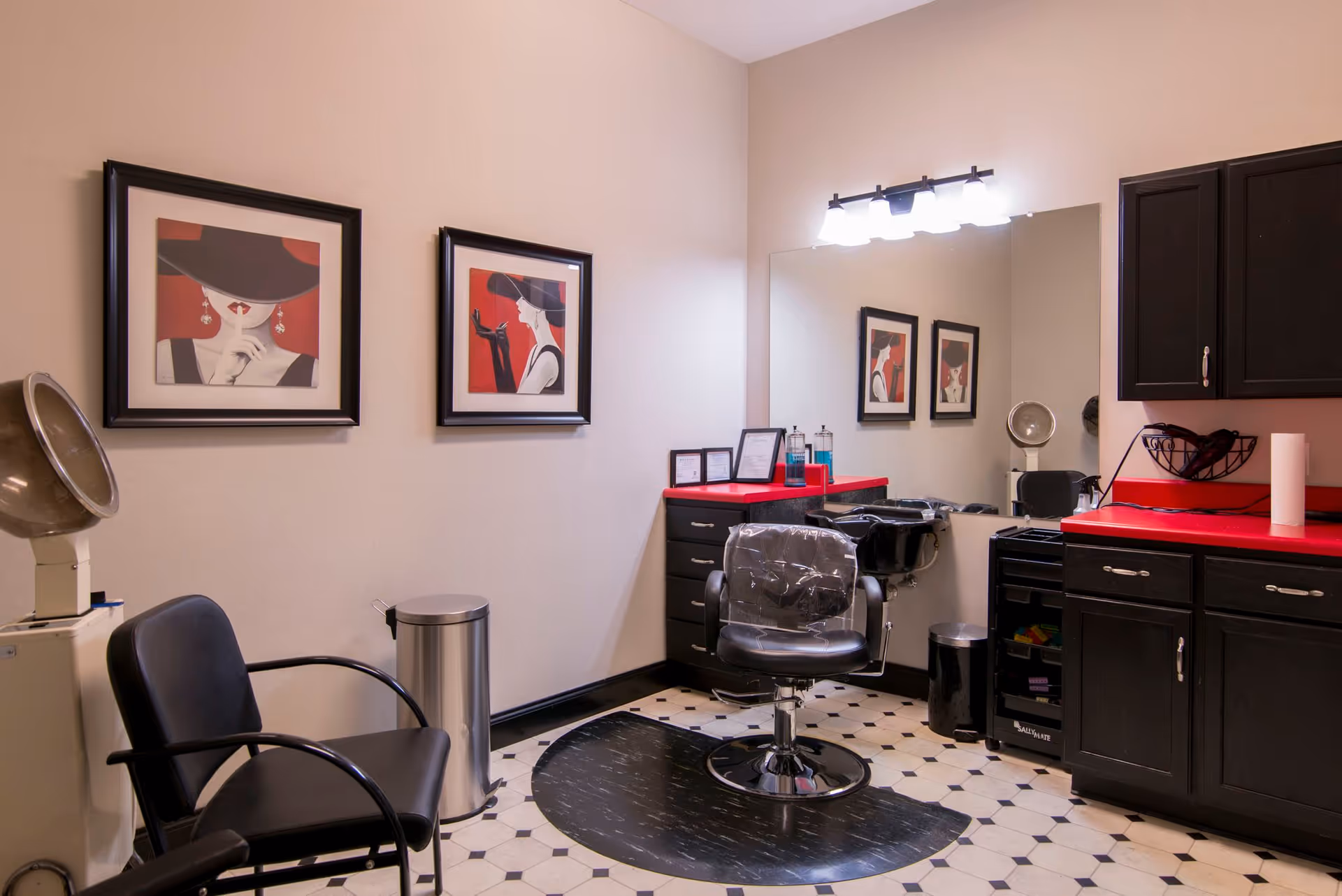 Interior view of a salon area in a senior living facility with a black salon chair covered in plastic, a black waiting chair, a hair dryer, black cabinets with a red countertop, a large mirror, and two framed artworks on the wall depicting women in black hats with red backgrounds.