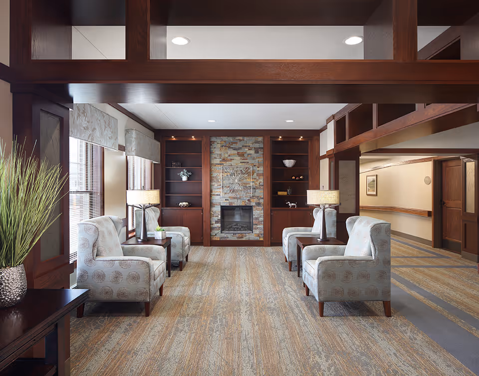 A cozy assisted living facility lounge area featuring four patterned armchairs arranged in pairs facing each other with side tables and lamps between them. The room has large windows with blinds and valances on the left, a stone fireplace with built-in wooden shelves on either side at the far end, and a carpeted floor with a striped pattern. The walls and ceiling are light-colored, and the wood trim and beams are dark brown, creating a warm and inviting atmosphere.