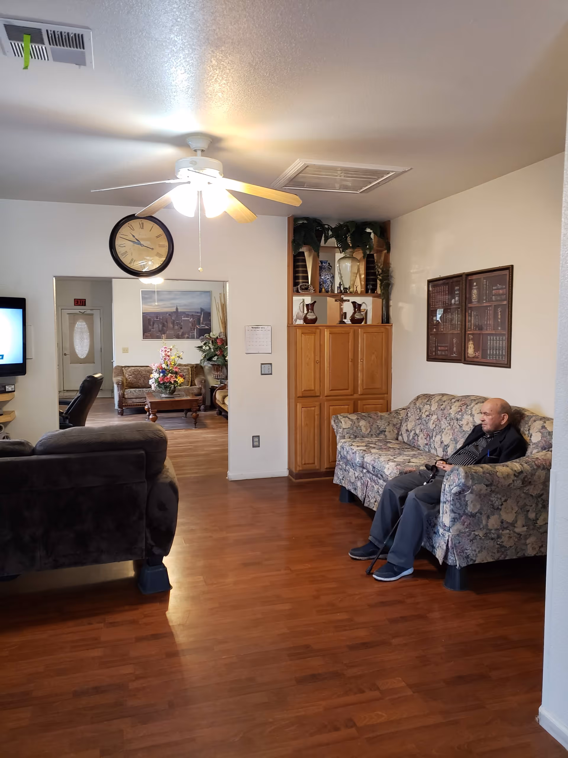A living room area in St. Luke's Residential Facility with wooden flooring, a ceiling fan with lights, a large wall clock, and a floral-patterned couch where an elderly man is seated holding a cane. There is a wooden cabinet with decorative items on top and a framed picture on the wall. In the background, another seating area with a floral arrangement on a coffee table is visible.