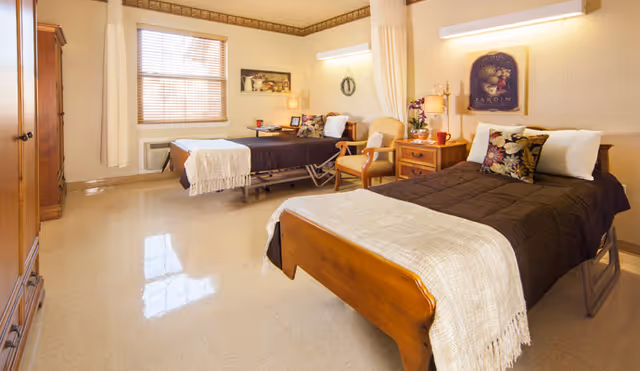 Well-lit shared patient bedroom with two single beds, bedside tables, chairs, and a window.