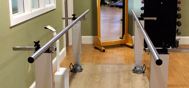 Physical therapy parallel bars in a rehabilitation center hallway with a wooden floor and green walls, a large mirror at the end, and exercise equipment visible on the right side.