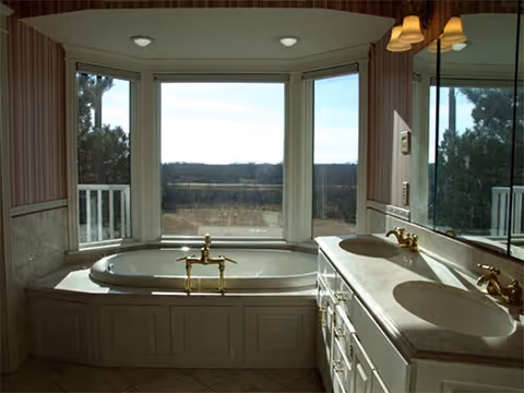 A bathroom featuring a large built-in bathtub with brass fixtures positioned in front of three large windows offering a view of the outdoors. To the right, there is a double sink vanity with brass faucets and a large mirror above it. The walls have striped wallpaper and the floor is tiled.