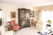 A bright living room with cream-colored walls and carpet. The room features a dark wooden china cabinet with glass doors, a red upholstered armchair with a side table and lamp, a beige armchair near a window with draped curtains, and a small wooden chair. There are framed pictures on the walls and a coffee table in the foreground.