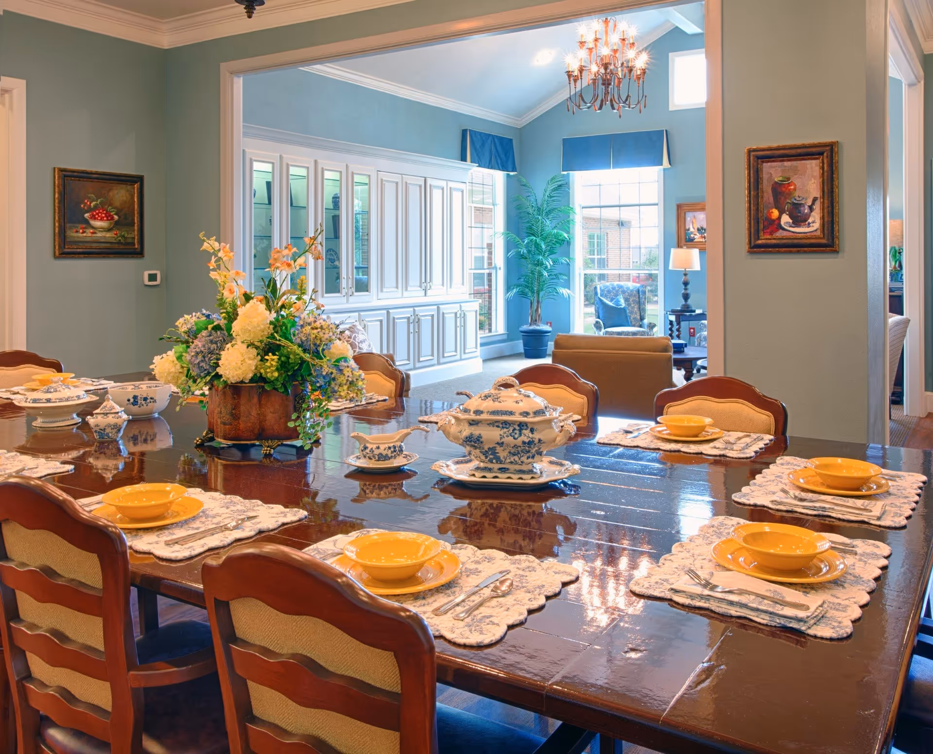 A formal dining room with a large polished wooden table set with yellow bowls, plates, and silverware on white lace placemats. A floral centerpiece decorates the middle of the table. The room has light blue walls, two framed paintings, and an adjacent living area with large windows, a potted plant, and a chandelier hanging from the ceiling.