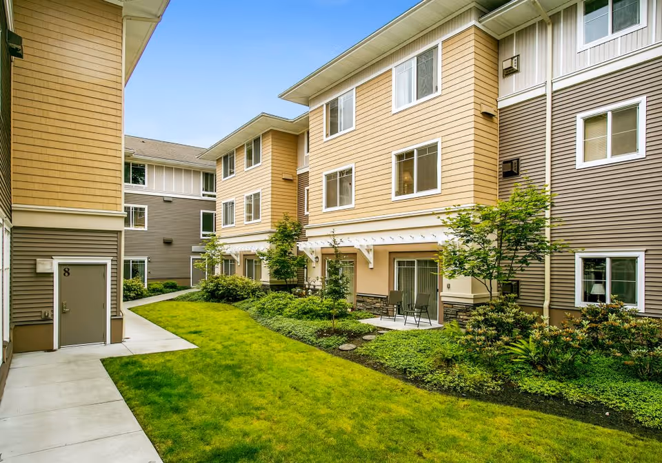 Exterior view of a senior living facility courtyard with well-maintained green grass, small trees, shrubs, and a concrete walkway. The building has beige and gray siding with multiple windows and a small patio area with chairs.