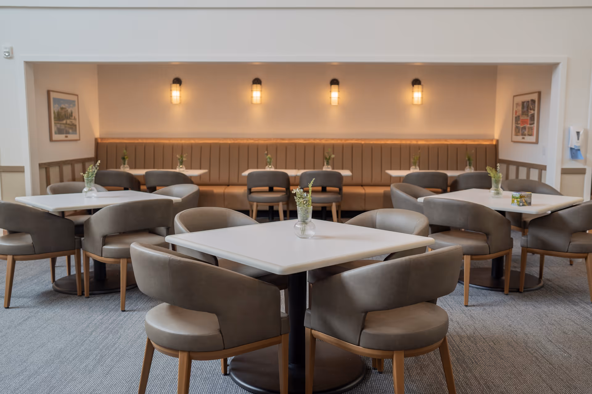 A dining area with multiple square tables surrounded by cushioned chairs. The back wall features a long upholstered bench with small vases of flowers on each table. Warm wall sconces provide lighting, and framed pictures hang on the side walls. The room has a calm and inviting atmosphere.