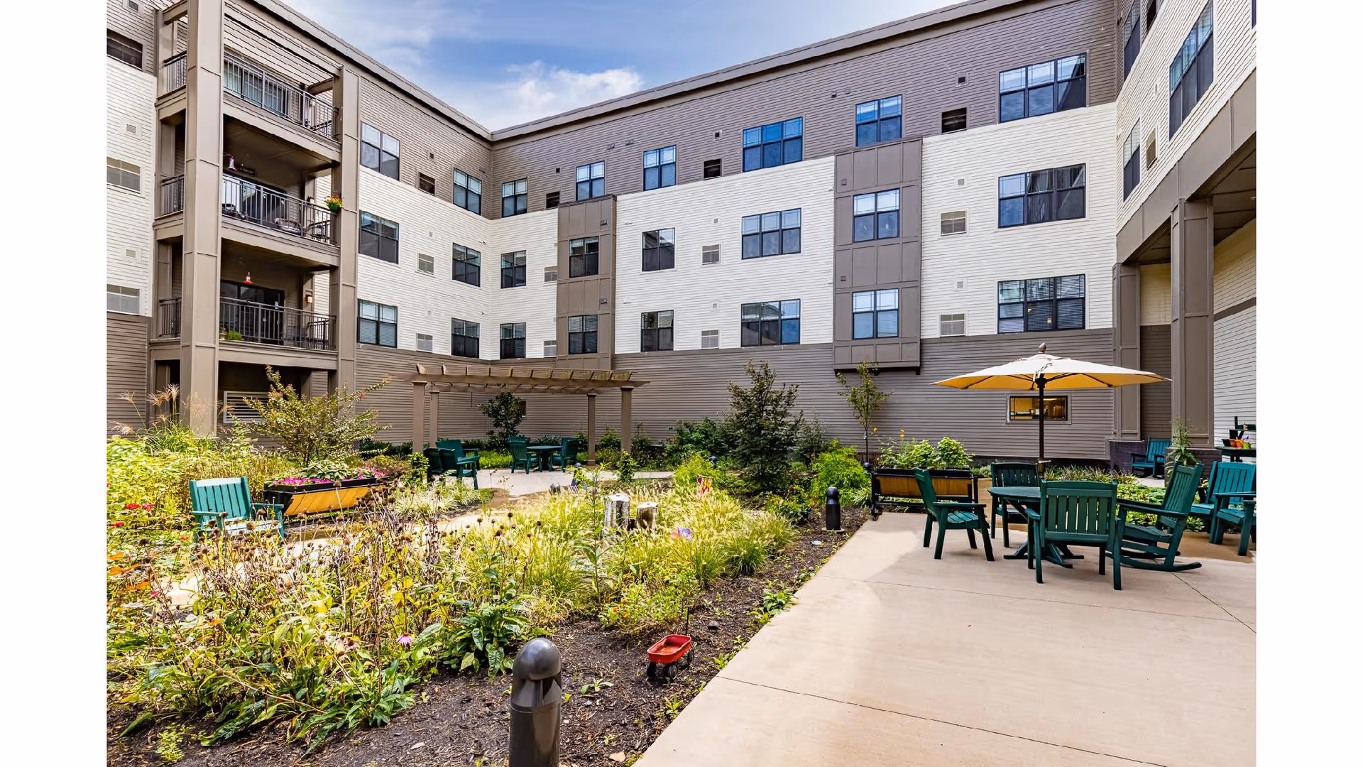 Outdoor courtyard area of a senior living facility with garden beds, green chairs, tables with umbrellas, and a pergola. The courtyard is surrounded by a multi-story building with many windows.