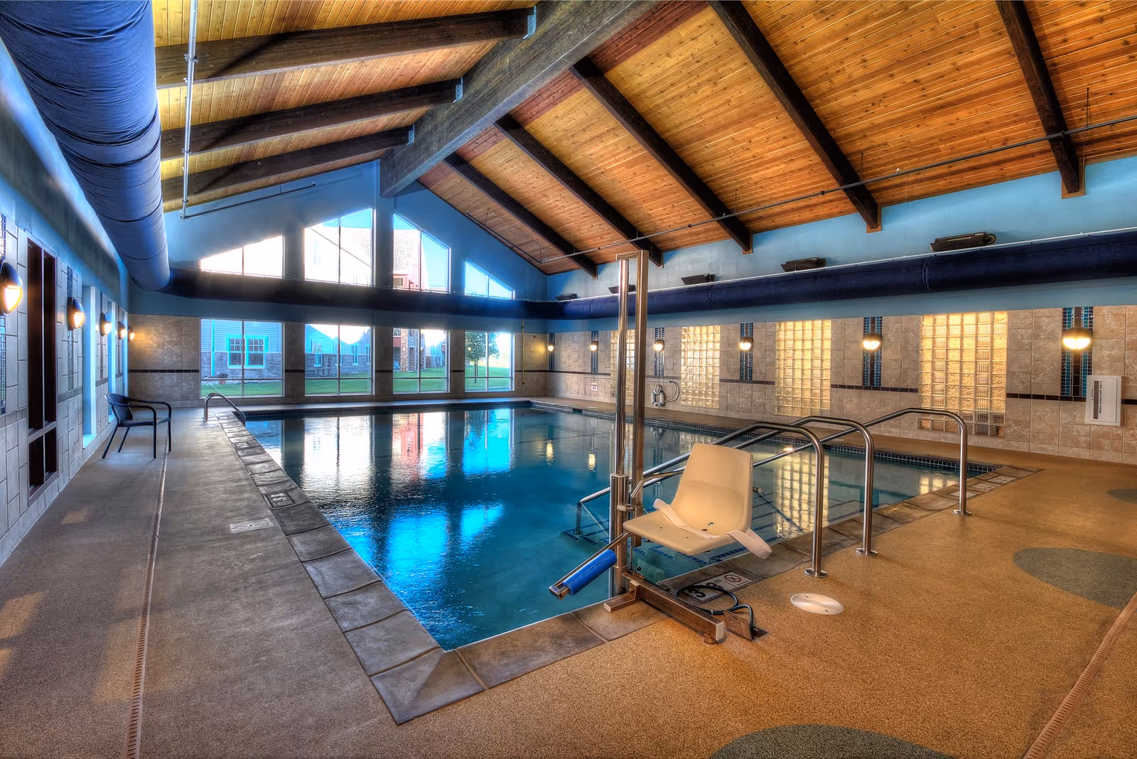 Indoor swimming pool with a wooden vaulted ceiling, pool lift, and large windows overlooking the outside.