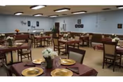 A dining room with multiple tables covered in maroon tablecloths and set with gold plates and floral centerpieces. The room has beige walls with framed pictures and decorations, and several chairs are arranged around each table.