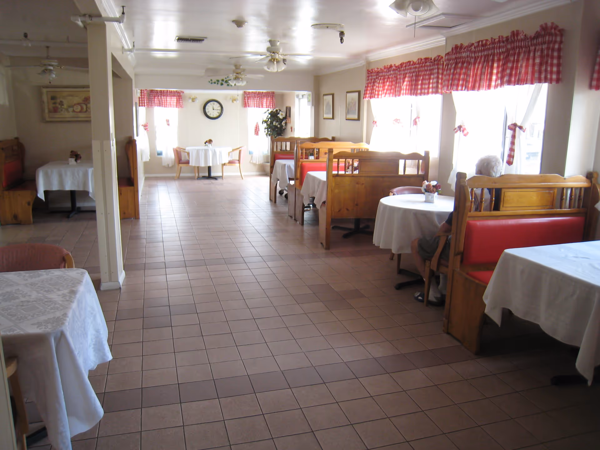 A dining room with tiled floor, wooden booths with red cushions, and tables covered with white tablecloths. The windows have white curtains with red checkered valances. A clock is mounted on the far wall, and a person is seated at one of the booths near the windows.