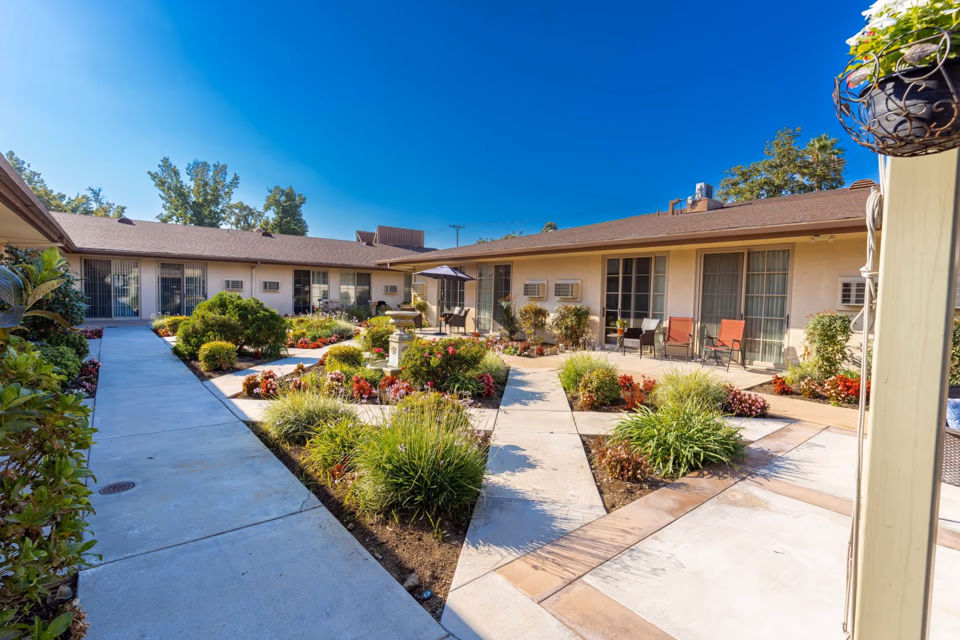 Outdoor courtyard area of a senior living facility with a clear blue sky, surrounded by single-story buildings with large windows and air conditioning units. The courtyard features well-maintained landscaping with various shrubs, flowers, and a central fountain. There are paved walkways and patio seating with chairs and an umbrella.