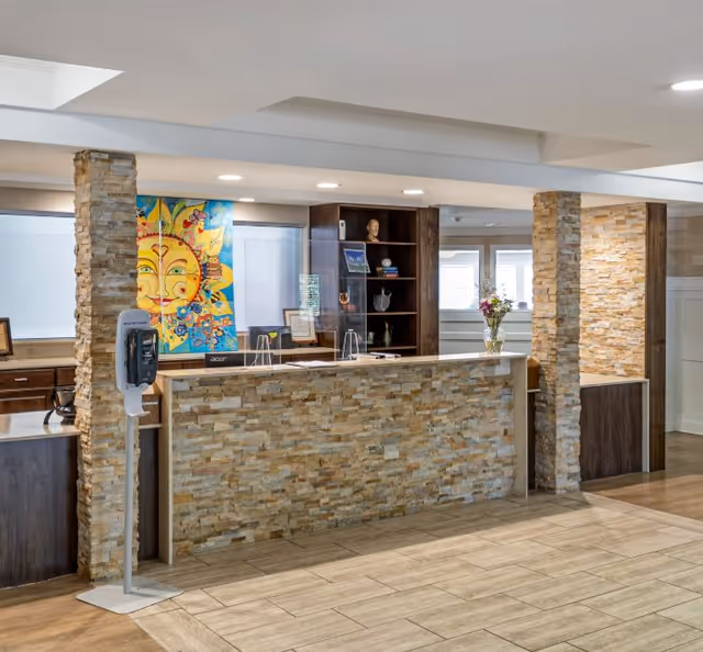Reception area with a stone-faced front desk, two stone pillars, a colorful sun-themed artwork on the wall, a hand sanitizer dispenser on a stand, and shelves with decorative items and flowers in a well-lit interior space.