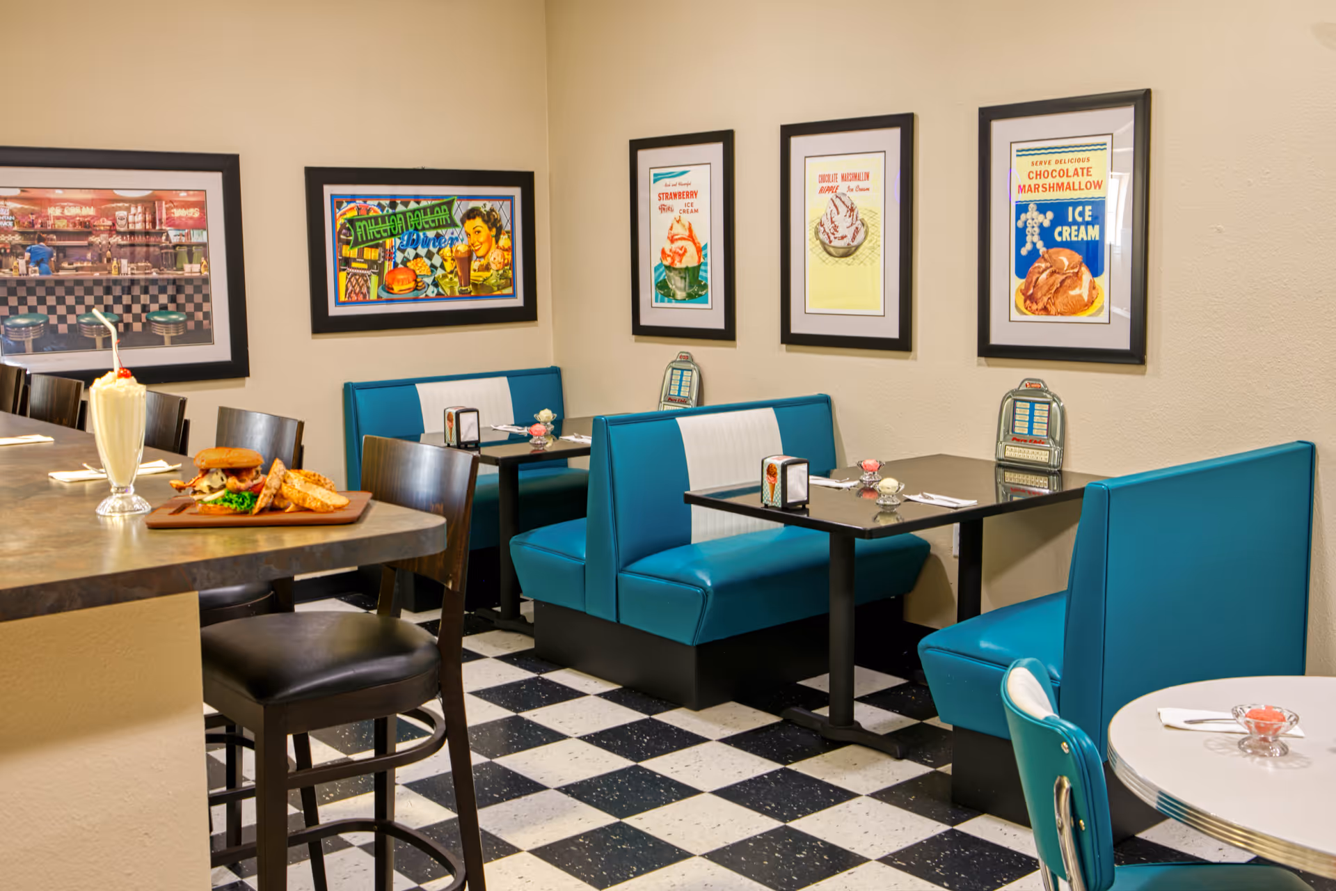 Interior view of a retro-style dining area with turquoise and white booth seating, black and white checkered floor, and framed vintage ice cream and diner-themed posters on the beige walls. A counter with black chairs is visible with a milkshake and a sandwich with fries on a wooden board.