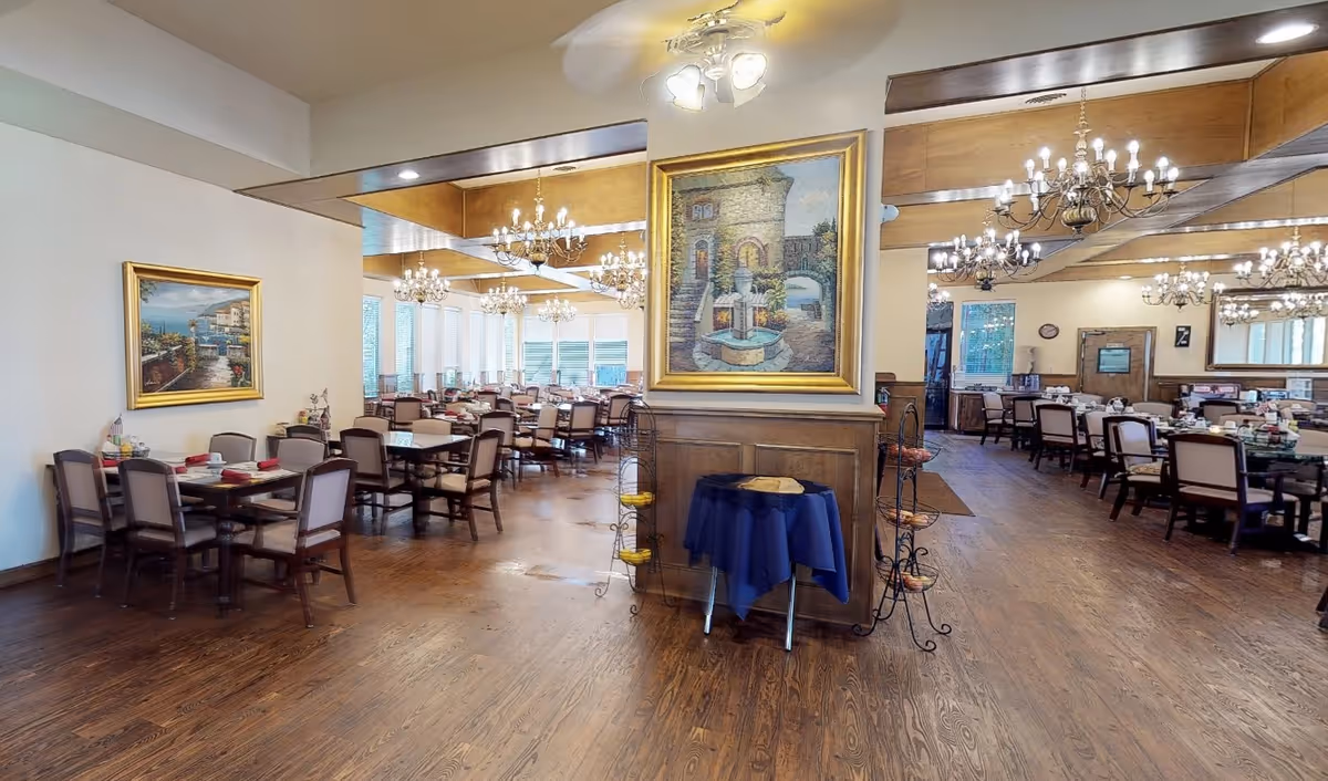 Spacious dining room in a senior living community with multiple tables and chairs arranged neatly. The room features wooden flooring, chandeliers hanging from the ceiling, and large framed paintings on the walls. There is a small table with a blue cloth and fruit baskets near a wooden partition in the center.