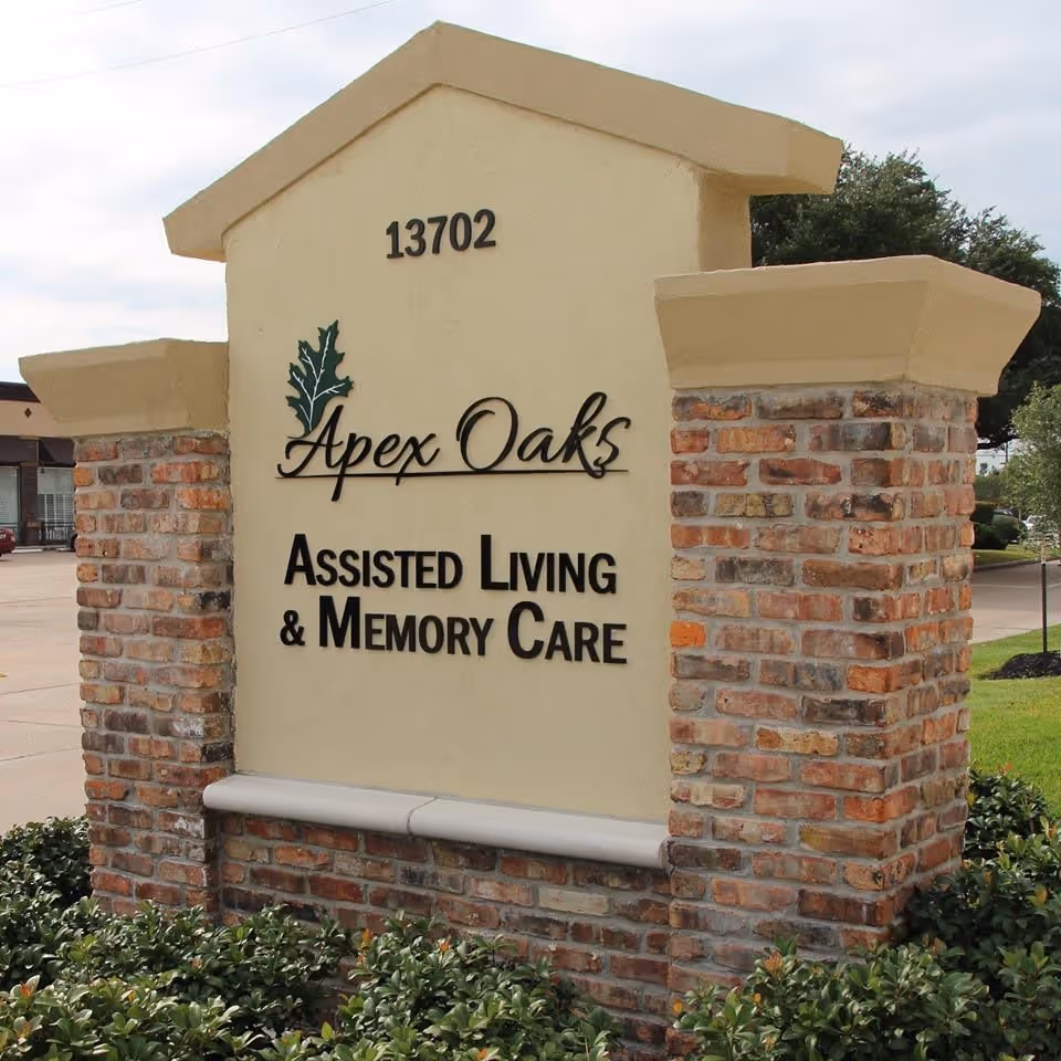A brick and stucco sign for Apex Oaks Assisted Living & Memory Care with the address number 13702 displayed at the top, surrounded by greenery and a parking lot in the background.