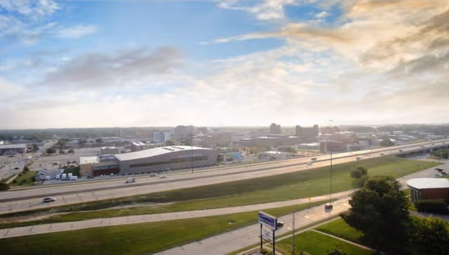 A wide aerial view of a cityscape with a highway running through the middle, surrounded by buildings, green grassy areas, and a partly cloudy sky during sunset or sunrise.
