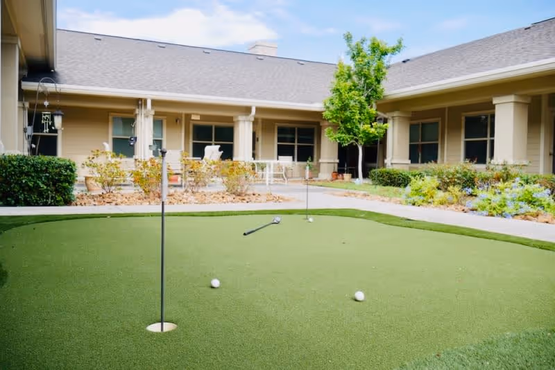 Outdoor putting green with golf balls and a golf club in the center of a courtyard surrounded by a single-story building with beige walls, windows, and a covered patio area with chairs and plants.