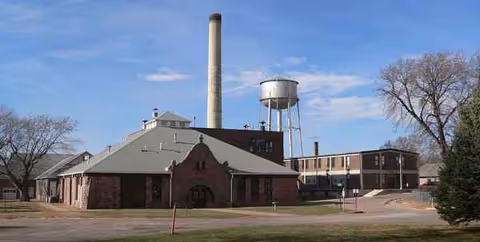 Brick facility with smokestack and water tower behind trees and a parking lot.
