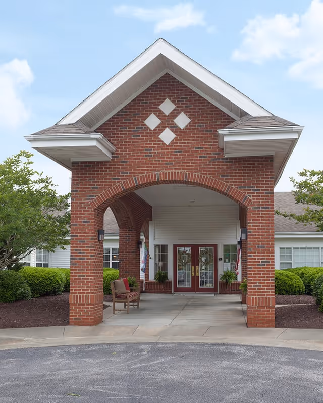 Entrance of a senior living facility with a covered brick archway, two red double doors, a bench, and greenery on either side under a partly cloudy sky.