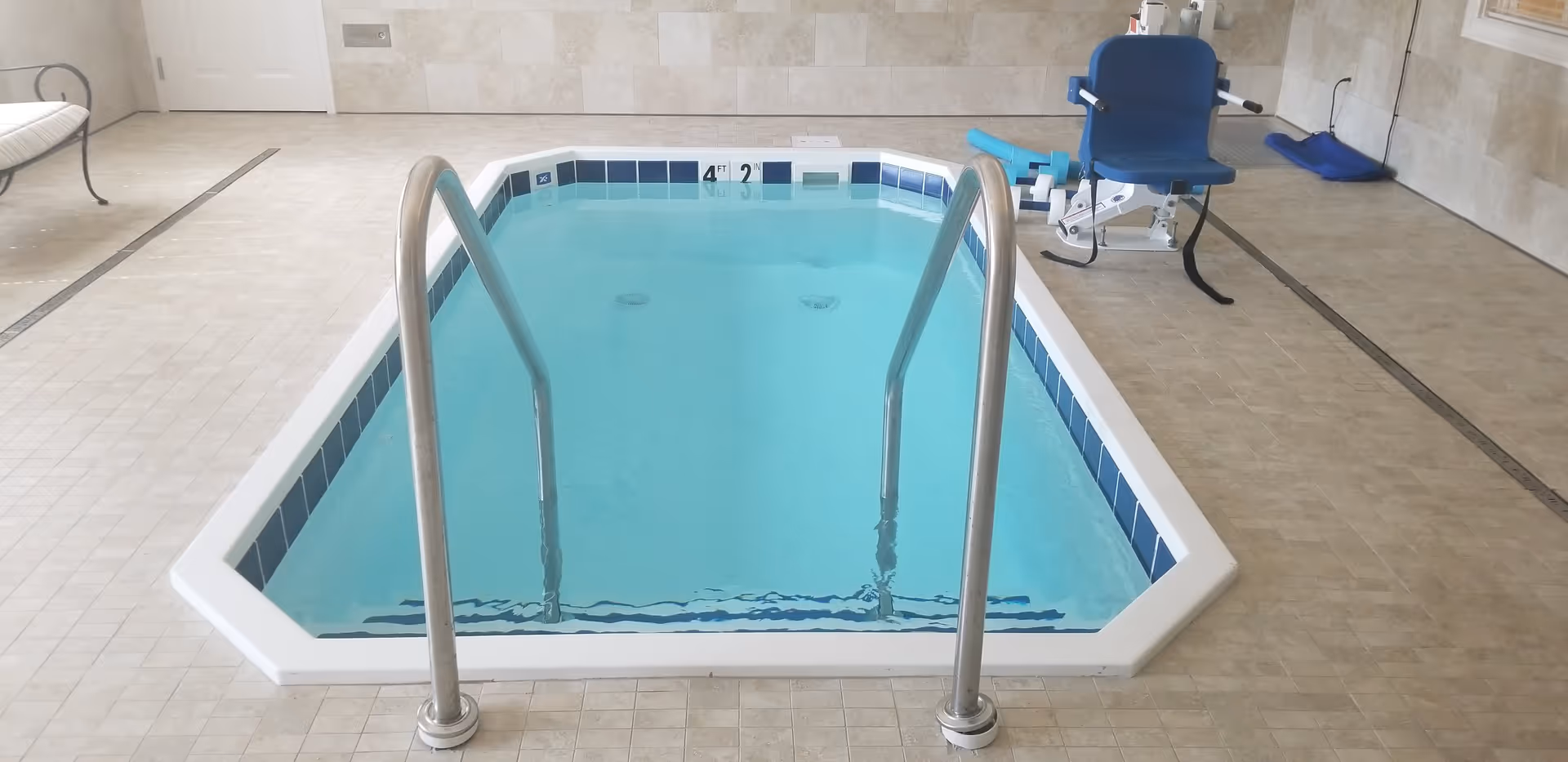 Indoor small rectangular pool with metal handrails on both sides for entry. The pool has blue tiles along the waterline and depth markers indicating 4 feet 2 inches. There is a blue pool lift chair on the right side for accessibility. The surrounding floor is tiled in beige, and a bench is visible on the left side against the wall.