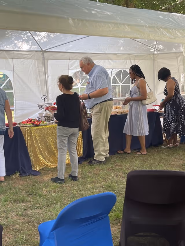 Several people standing in line under a white outdoor canopy tent, serving themselves food from tables covered with black and gold tablecloths. The setting appears to be a garden or grassy outdoor area with chairs in the foreground.