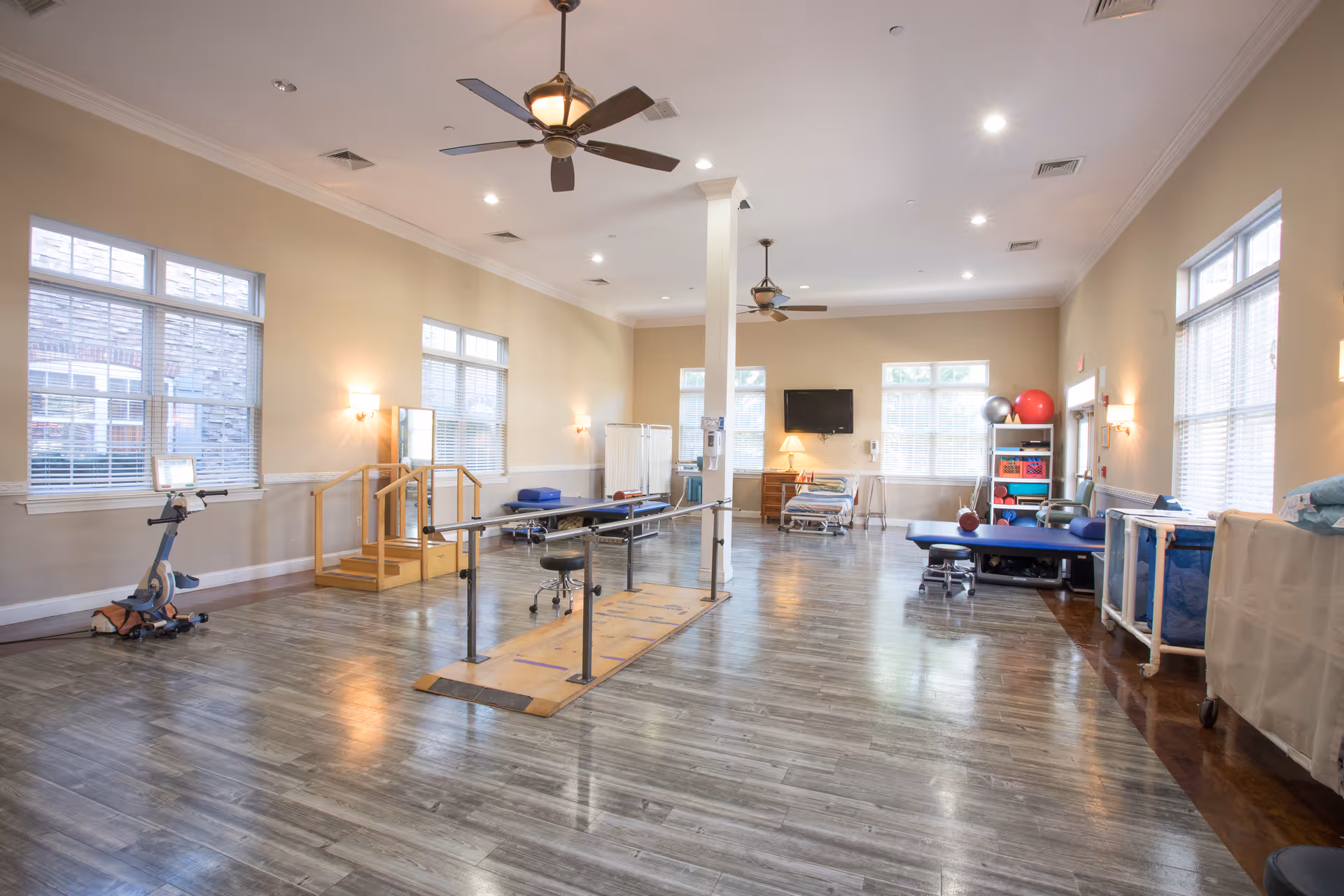 A spacious rehabilitation or physical therapy room with wooden flooring, parallel bars for walking exercises, exercise equipment including a stationary bike, therapy tables, exercise balls, and a TV mounted on the wall. The room has large windows allowing natural light, ceiling fans, and soft wall lighting.