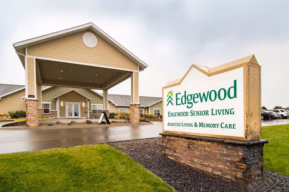 Exterior view of Edgewood Senior Living facility showing the entrance with a covered driveway and a large sign in the foreground that reads 'Edgewood Senior Living Assisted Living & Memory Care'. The building is beige with stone accents and there are parked cars visible in the background.