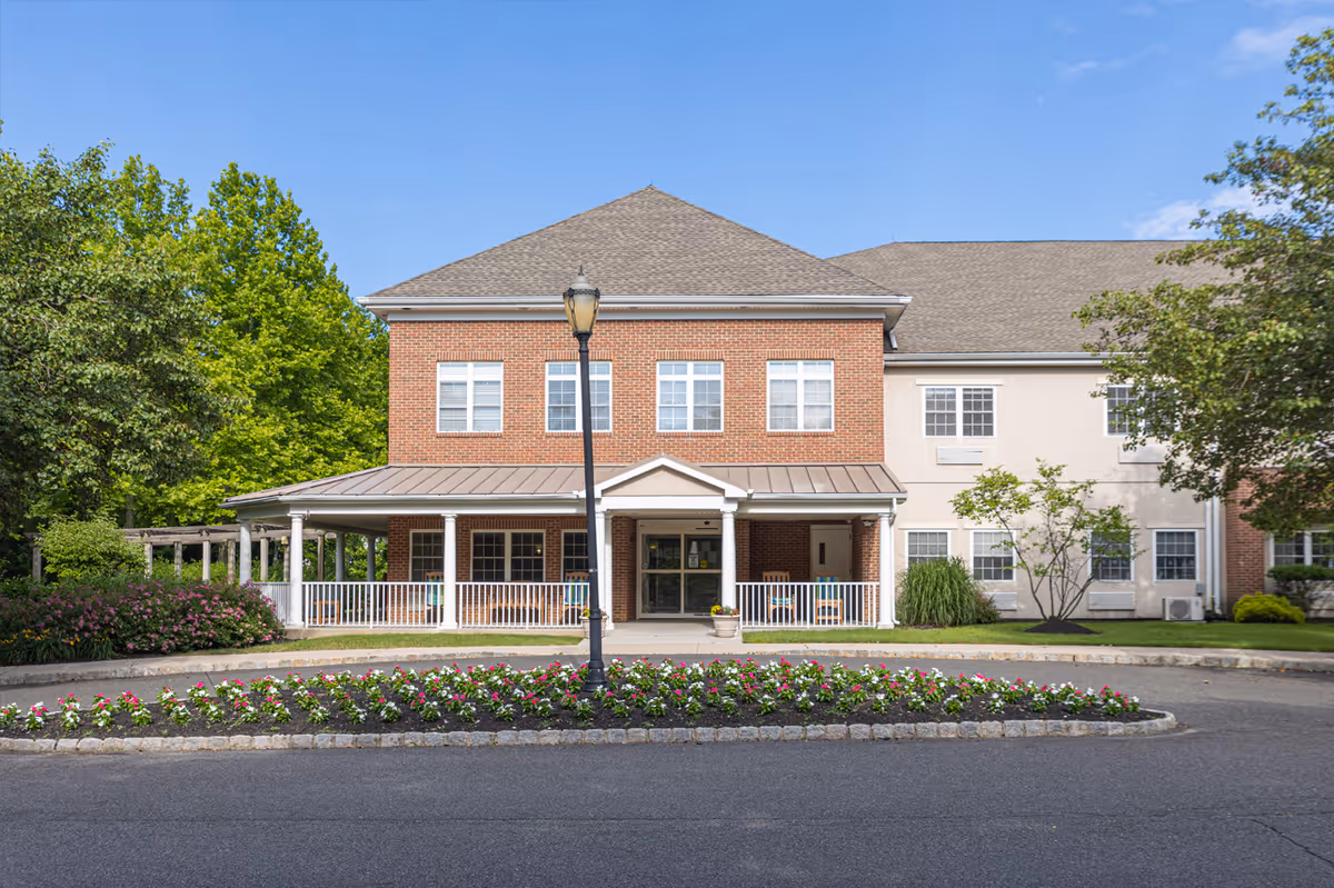 Front exterior view of a two-story senior living facility building with a brick and light-colored facade, a covered entrance porch with white columns, a lamp post in front, and a flower bed with colorful flowers along the driveway. Trees and shrubs surround the building under a clear blue sky.