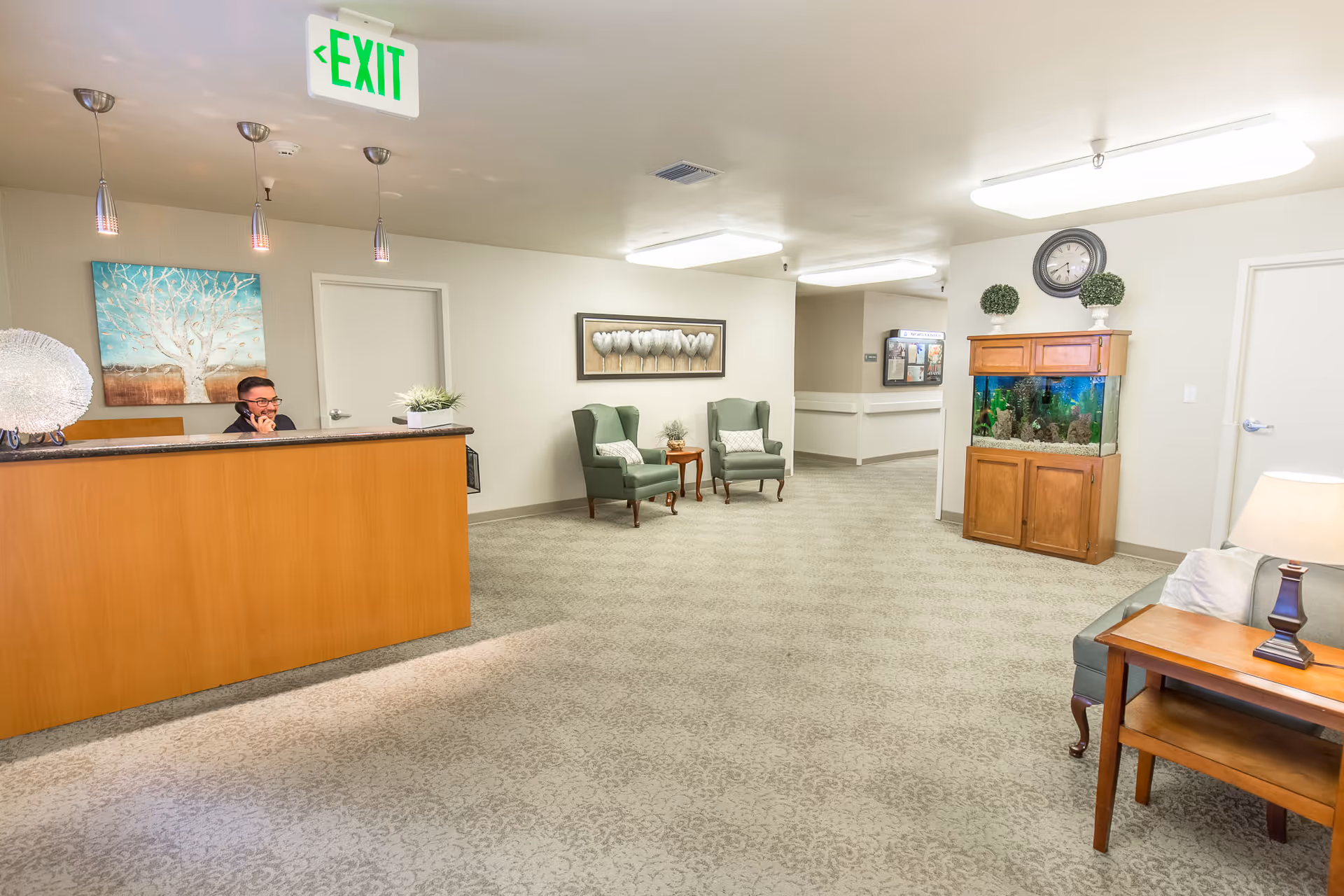 Reception area of Windsor Monterey Care Center with a receptionist sitting behind a wooden desk. The room features two green armchairs with a small table between them, a large fish tank on a wooden cabinet, a wall clock, and decorative plants. The walls are light-colored and there is a painting of a tree behind the desk. The carpet has a subtle pattern, and there is an illuminated exit sign on the ceiling.