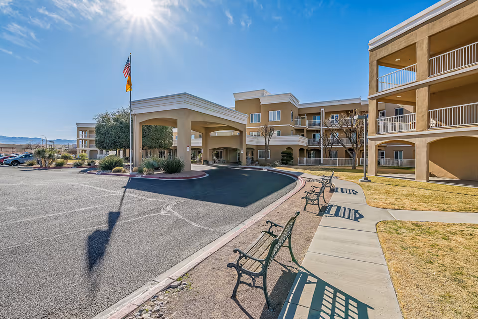 Exterior view of Solstice Senior Living at Las Cruces showing a three-story building with balconies, a covered entrance with two flagpoles displaying the American and New Mexico flags, a parking lot, and several benches along a sidewalk in a landscaped area with grass and trees under a clear blue sky.