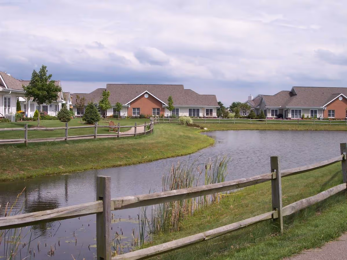A peaceful outdoor scene at Wesleyan Meadows featuring a small pond bordered by a wooden fence, with well-maintained grassy areas and several single-story residential buildings in the background under a cloudy sky.