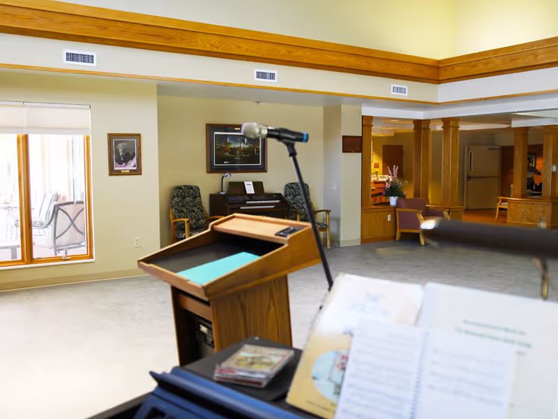 Wooden podium and microphone set up in a bright common room with a piano, chairs, and columns.