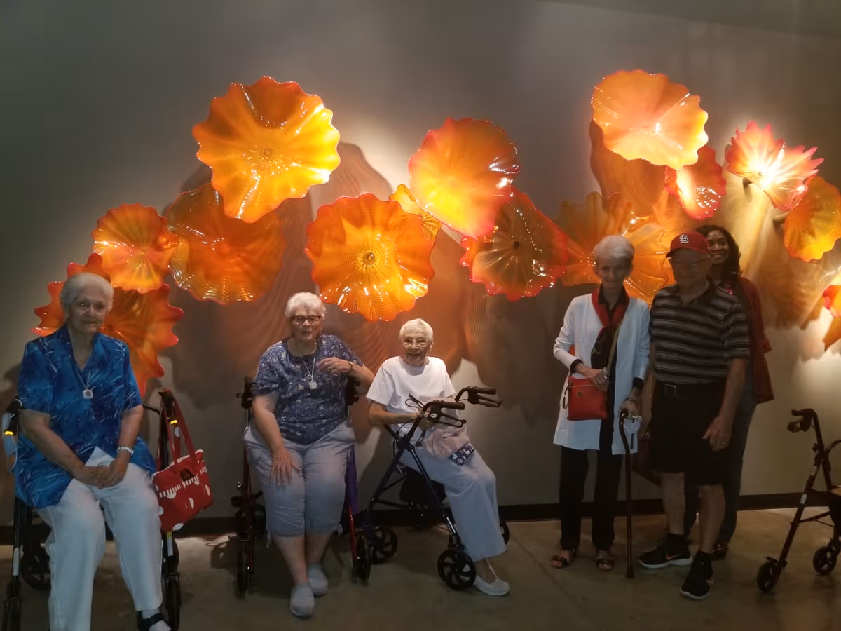 A group of elderly people, three seated with walkers and two standing, posing in front of a wall decorated with large, bright orange and yellow glass art pieces shaped like flowers or sea creatures.