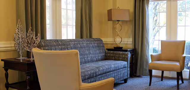 A cozy living room area with a patterned blue sofa, two beige armchairs, a wooden side table with decorative metal leaf sculptures, a table lamp with a beige shade, and large windows with green curtains letting in natural light.