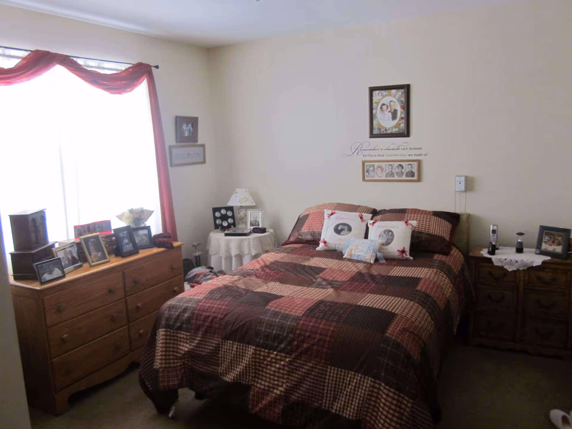 A cozy bedroom with a bed covered in a patchwork quilt in shades of brown and red. The bed has three decorative pillows with photos and the word 'Memories' on them. On the left side of the bed is a wooden dresser topped with multiple framed photographs and a small lamp. To the right of the bed is a wooden nightstand with a lace doily, a framed photo, and a cordless phone. The walls are light-colored and decorated with framed pictures and a wall decal that reads 'Remember to cherish each moment for this is what memories are made of.' A window with sheer curtains and a red valance lets in natural light.