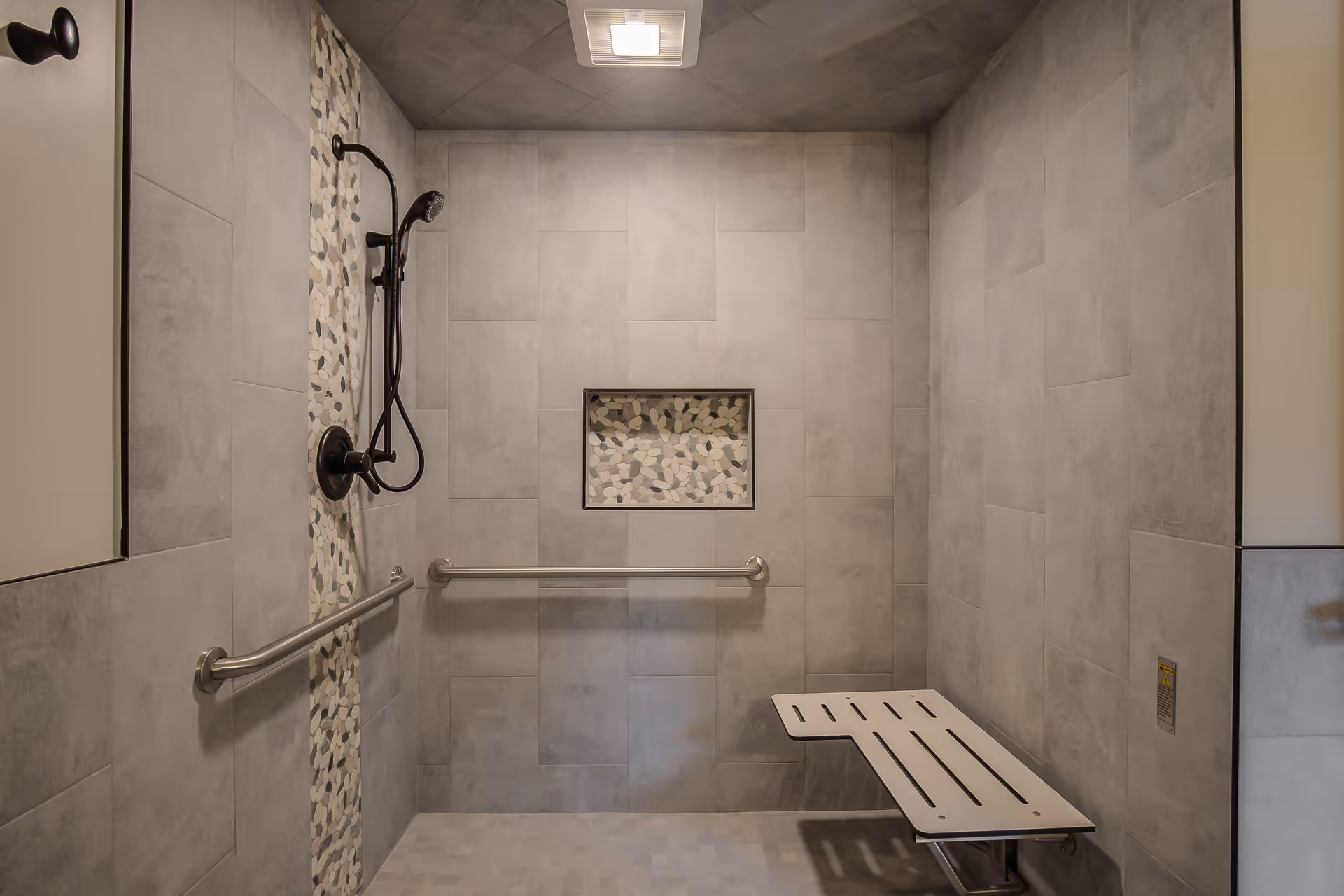 A modern accessible shower area with gray tiled walls and floor, a vertical strip of pebble-like decorative tiles behind the black showerhead and controls, a horizontal grab bar on the back wall, and a foldable white shower bench attached to the right wall.