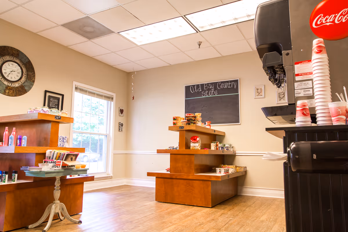 Interior view of a small store area inside a facility with wooden shelves displaying various snacks, toiletries, and other small items. A Coca-Cola soda fountain machine with stacked cups is visible on the right. A chalkboard on the wall reads 'Old Bay Country Store'. There is a window letting in natural light and a decorative clock on the wall.