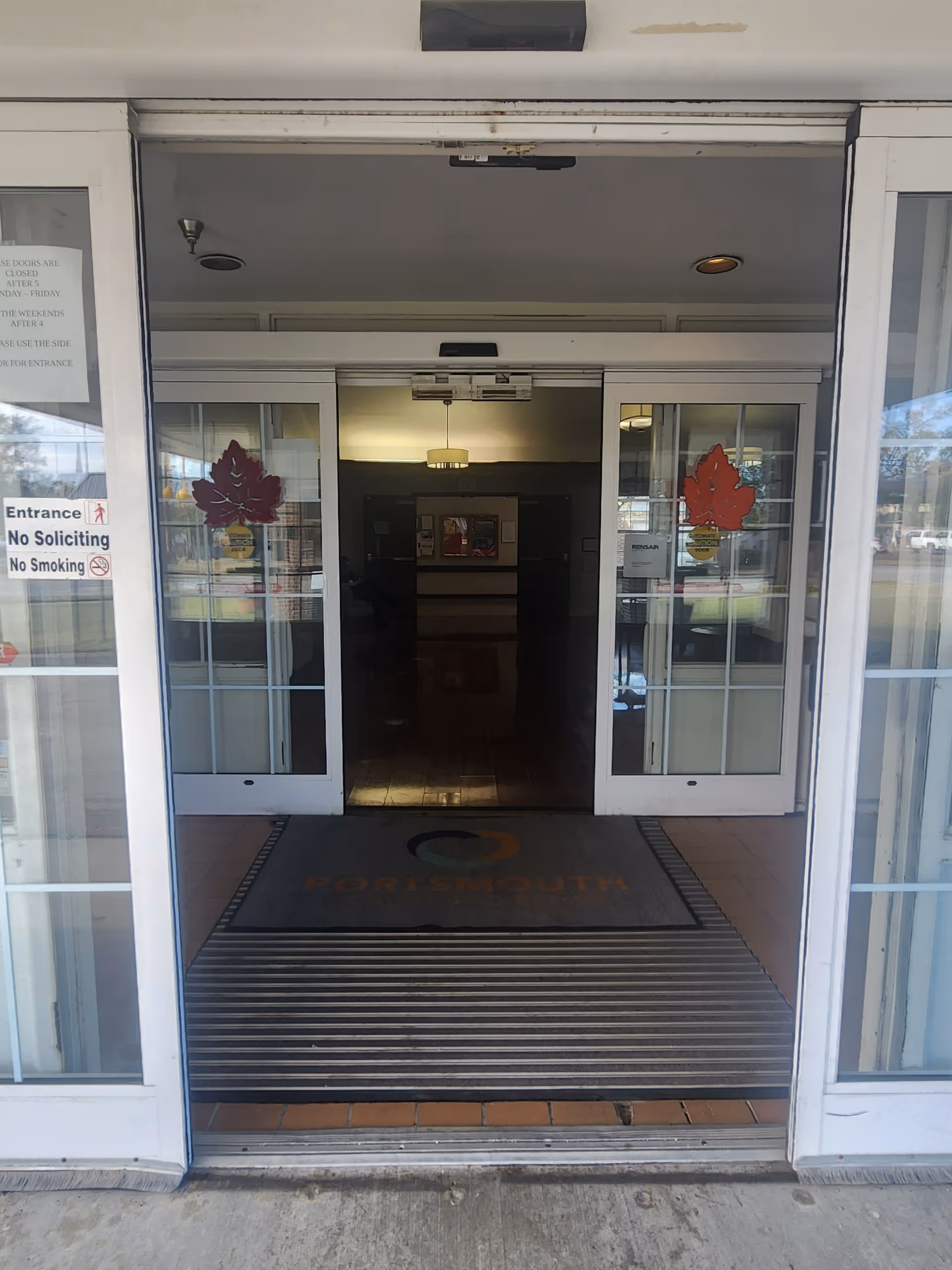 Automatic sliding glass entry doors open to the Portsmouth lobby, showing a doormat with "Portsmouth", maple leaf decals on the glass, and interior lighting.