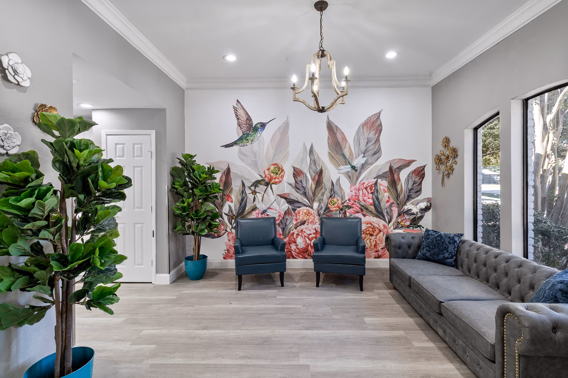 A bright and inviting living room area with a large floral and hummingbird mural on the back wall. The room features two blue armchairs, a long gray tufted sofa with blue cushions, two large potted plants, and a chandelier hanging from the ceiling. Large windows on the right side allow natural light to fill the space.