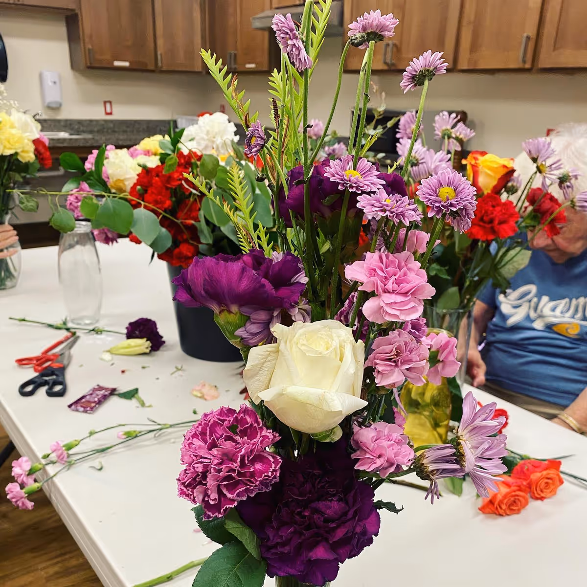 A colorful bouquet of roses, carnations, and daisies sits on a table amid floral-arranging supplies in a senior living activity room.