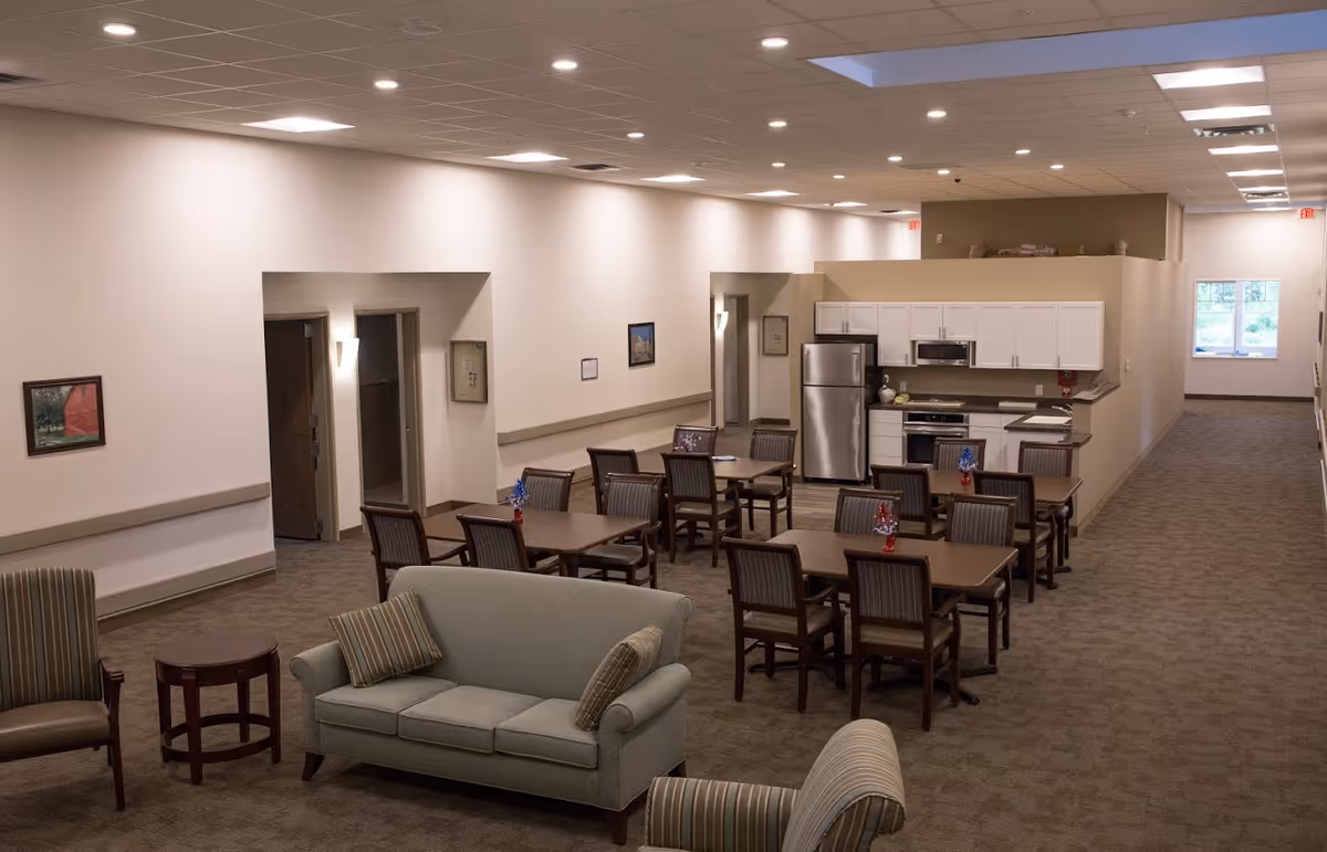 Interior view of a senior living facility common area featuring a small kitchen with white cabinets and stainless steel appliances, several wooden tables with chairs arranged for dining or activities, and a seating area with a sofa and armchairs. The space is well-lit with ceiling lights and has neutral-colored walls and carpet.