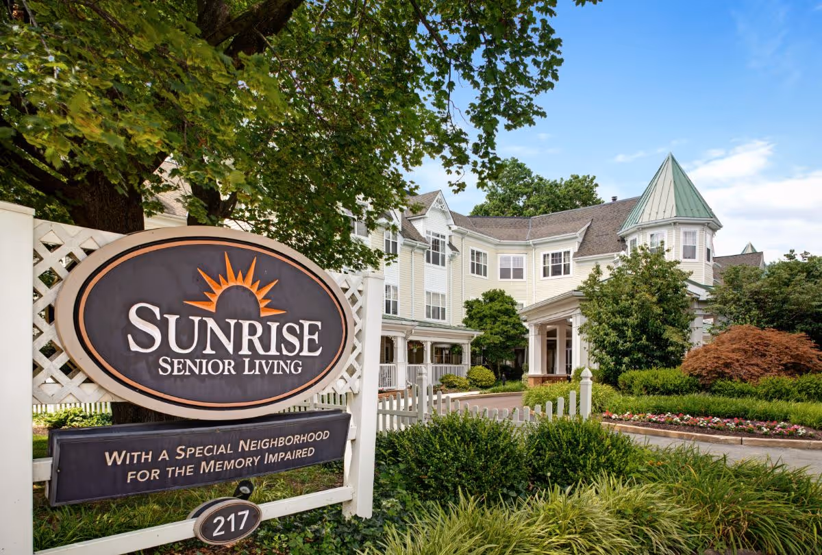 Exterior view of Sunrise Senior Living facility with a large sign in the foreground that reads 'Sunrise Senior Living - With a Special Neighborhood for the Memory Impaired'. The building is a multi-story structure with light-colored siding, a green roof turret, surrounded by trees, bushes, and a flower bed under a blue sky.