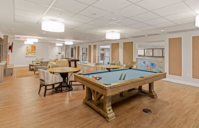 A spacious and well-lit common area with a pool table in the foreground, surrounded by several round tables with chairs. The room features wood flooring, modern ceiling lights, and decorative wall panels. There is a seating area with sofas and a painting on the wall in the background.
