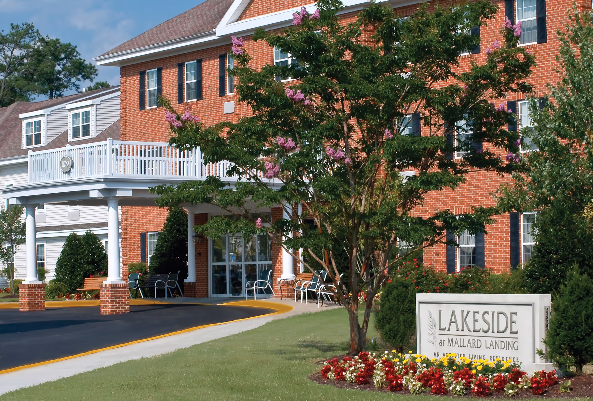 Exterior view of a senior living facility named Lakeside at Mallard Landing, featuring a red brick building with white columns and a covered entrance. There is a tree with pink flowers and a landscaped flower bed in front of the building, along with a sign displaying the facility's name.