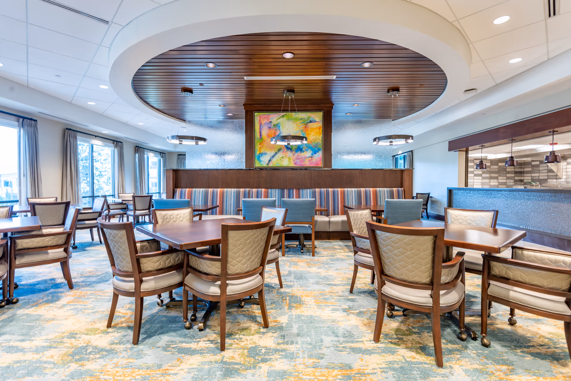 A bright and spacious dining room in a retirement community featuring multiple wooden tables with cushioned chairs arranged neatly. The room has large windows with curtains allowing natural light to fill the space. A circular wooden ceiling feature with modern pendant lights hangs above a booth seating area with striped upholstery and a colorful abstract painting on the wall behind it. The floor is covered with a patterned carpet in shades of blue and beige.