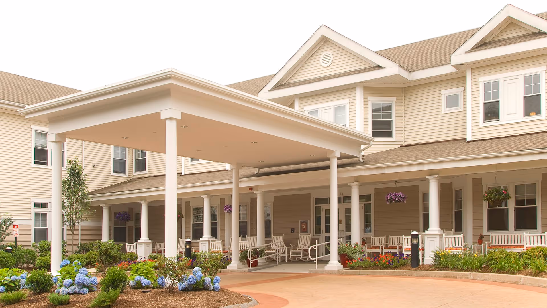 Exterior view of Atria Longmeadow Place senior living facility showing a covered entrance with white pillars, surrounded by landscaped gardens with blooming flowers and shrubs. The building has beige siding and multiple windows, with white rocking chairs arranged on the porch.