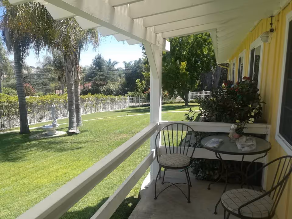 A covered porch area with two metal chairs with cushions and a glass-top table. The porch overlooks a well-maintained green lawn with palm trees, a white fence, and a small fountain. The building exterior is yellow with white trim and windows, and there are bushes with flowers near the porch.